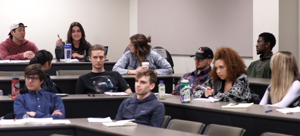 A group of students, one points her pen towards the front of the room