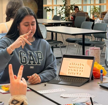 A student teaching sign language to peers, using a tablet showing hand signs as a visual aid
